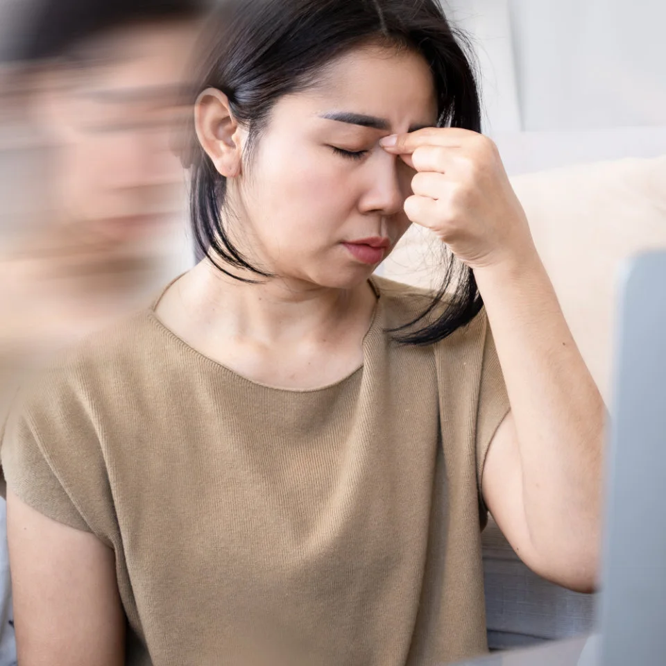 A woman sits with her eyes closed, pinching the bridge of her nose, appearing stressed or fatigued. A blurred duplicate image suggests dizziness or mental strain.