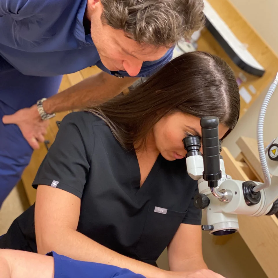 A woman in black scrubs looks through a microscope while a man in blue scrubs stands beside her, observing.