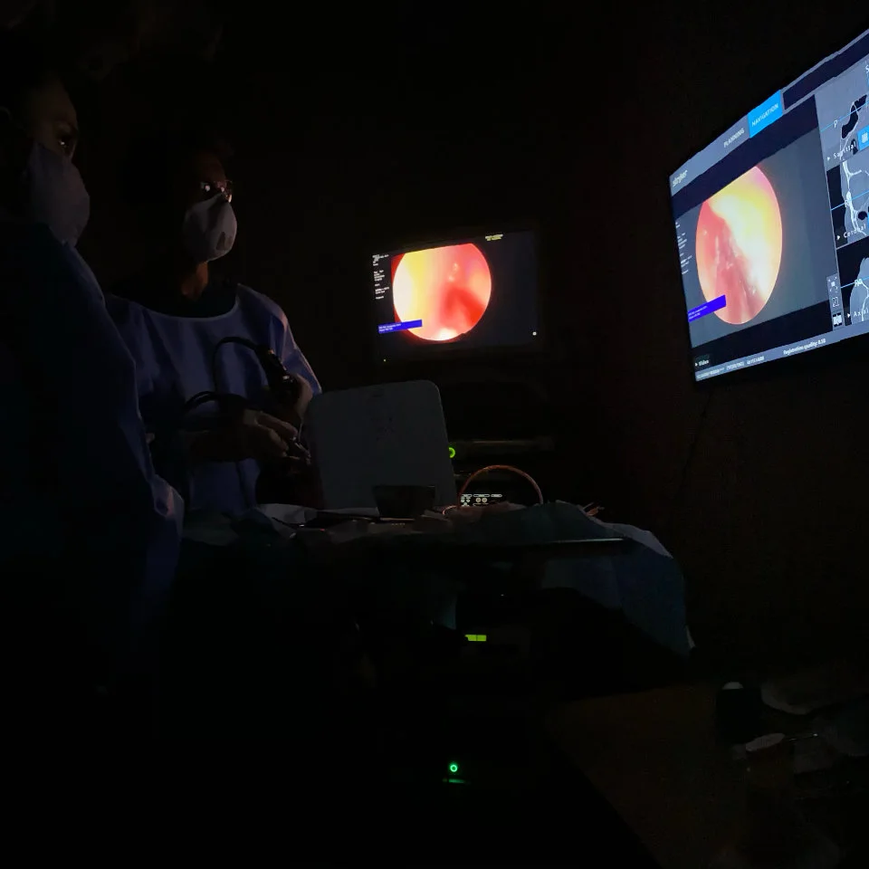 Two medical professionals in masks perform an endoscopic procedure in a dark room, monitoring images of an internal organ displayed on screens.