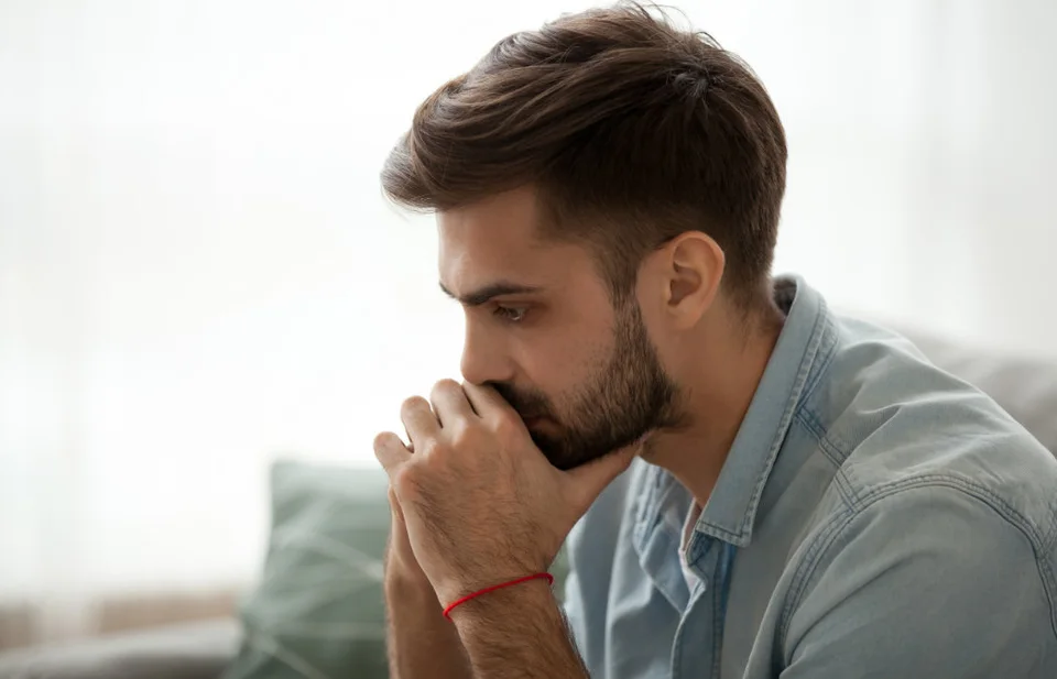 A man with a beard sits indoors, leaning forward with his hands clasped near his mouth, appearing deep in thought.