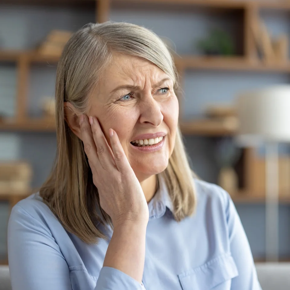 Elderly Caucasian woman suffering from otitis, touching her ear in pain, sitting in a modern living room. She looks distressed and worried about her earache.