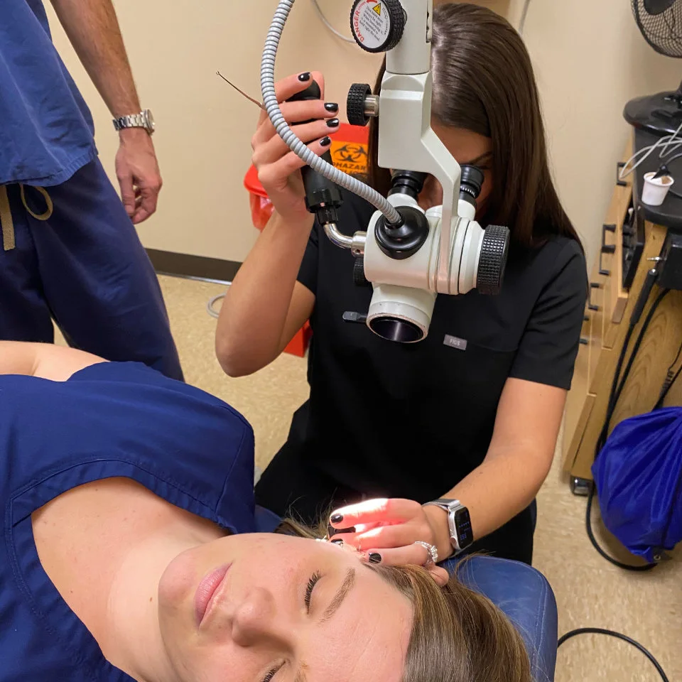 A healthcare professional examines a patient’s ear with a microscope and light while the patient lies on an exam chair.