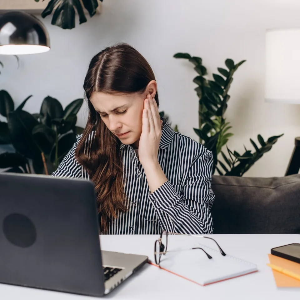 Upset young brunette female sit on sofa at table working laptop feeling ear pain, unhappy irritated businesswoman or student. Furuncle, earache, shingles, eczema, ear disease, otitis concept