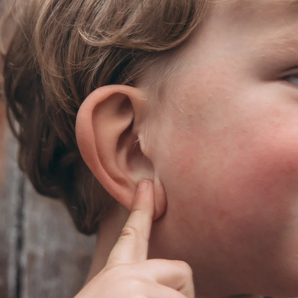Close-up of a child with light brown hair touching their ear with their index finger.